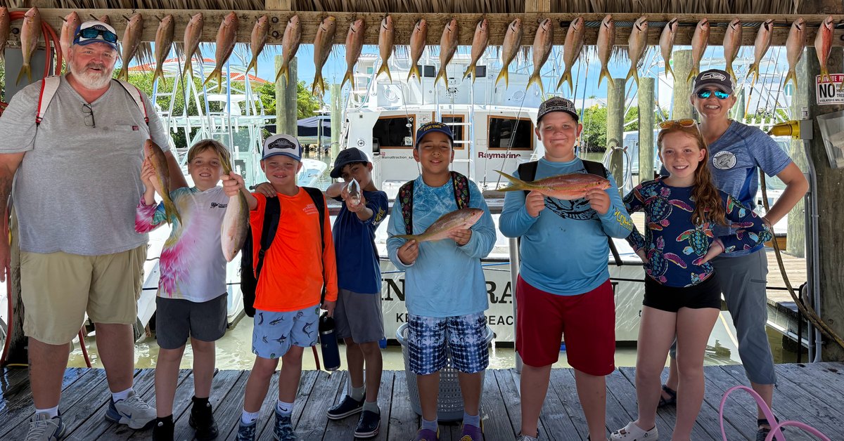 Stanley Switlik Elementary fishing club students holding their yellowtail snapper catch with chaperones under the tiki at Marathon Sailfish Marina, with a row of yellowtail hanging overhead